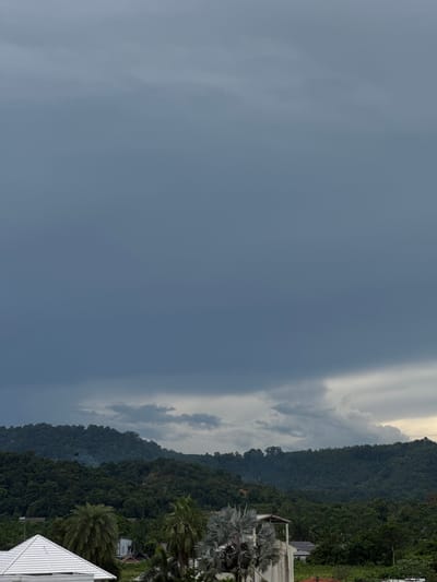 An overcast sky looms over a dense, lush green mountain range in Kathu, Thailand. In the foreground, a white tiled roof of a building is visible on the left, partially obscured by several palm trees. A concrete structure with a simple roof stands on the right, also surrounded by vegetation. The overall impression is one of a humid, tropical environment under a heavy sky, possibly indicating approaching rain.