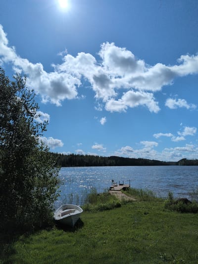 The image depicts a serene lakeside scene in Hartola, Finland, on a bright, sunny day. The upper half of the frame is dominated by a clear blue sky with scattered white cumulus clouds; the sun is intensely bright in the upper left, causing a lens flare. A dense line of dark green trees forms the distant horizon across the lake. In the immediate foreground on the left, a large deciduous tree with green foliage partially shades the grassy ground. The middle ground features a calm blue lake, its surface shimmering with numerous specular reflections from the sunlight. A small, white, wooden rowboat is pulled ashore on the green grass in the lower-left quadrant, positioned near the water's edge. To the right of the boat, a narrow wooden pier extends into the lake from the grassy shore, featuring a simple wooden chair facing out over the water. Green grass and reeds grow along the shoreline. No individuals are present in the scene.