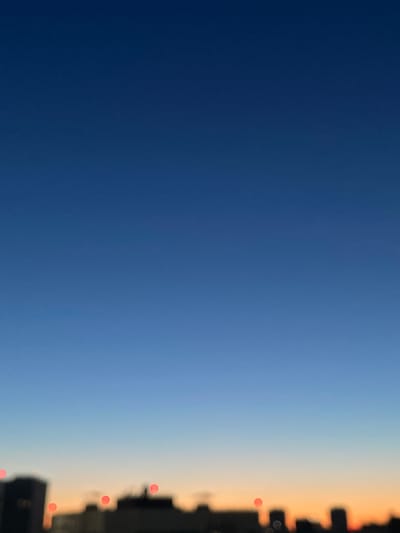 The image depicts a wide, unobstructed view of the sky above the city of Sinyavino, Russia, during twilight. The sky transitions from a deep, dark blue at the top to a lighter gradient of blue and orange near the horizon. The lower portion of the image features a blurred, silhouetted cityscape, indicating the presence of buildings. Several out-of-focus red circular lights are visible scattered across the skyline, likely streetlights or illuminated windows. No people or specific activities are discernible due to the blur and focus on the sky and distant cityscape.