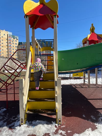 A child in a colorful jacket and gray pants climbs the yellow steps of a playground structure. The playground is located in Dobryanka, Russia, with a multi-story residential building visible in the background. Snow patches are present on the ground next to the brown playground surface. The playground structure features a green slide and red and yellow decorative elements.