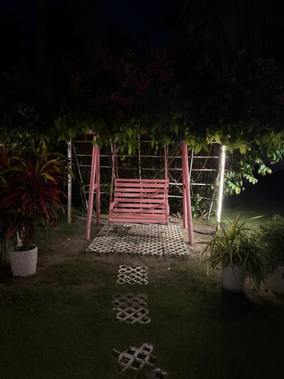 A pink swing set with a wooden seat is positioned in a garden at night. The swing is surrounded by lush green foliage and decorative plants in white pots. A pathway of decorative concrete pavers leads to the swing, and a vertical light fixture illuminates the scene. The background features a trellis structure and more greenery. The setting appears to be outdoors, possibly a residential garden or a public park area in Cam Lâm, Vietnam. There are no people or animals present.