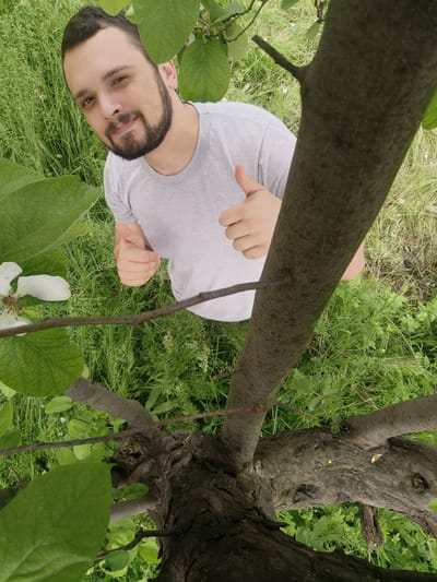 A man with a beard, wearing a gray t-shirt, is giving two thumbs up. He is standing in a grassy area, likely a field or yard, as indicated by the tall green grass surrounding him. In the foreground is a tree trunk with leaves and a single white flower. The location may be Popintsi, Bulgaria, though no landmarks are visible in the image to confirm the location. The camera is angled upwards, resulting in a perspective from below the man and the tree.