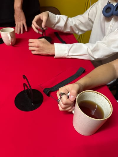 Two individuals are gathered around a table covered with a vibrant red cloth. On the left side of the table, a light-colored mug rests near the edge. In the center, a person's hands are occupied with a black object, seemingly manipulating strings or cords attached to it. To the right, another person's arm is visible, holding a mug filled with a dark liquid, possibly tea. This individual is wearing a white button-up shirt and a pair of blue headphones around their neck. The background is blurred but suggests an indoor setting with yellow and dark walls. There is no indication of the specific location within Chiang Mai City Municipality, Thailand.