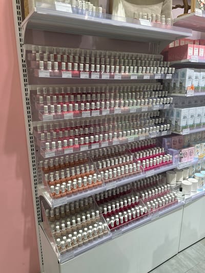 This is a high-angle, indoor shot of a retail display filled with numerous small bottles of what appear to be perfumes or essences. The bottles are neatly arranged in clear plastic shelving units, organized in rows and columns. The liquids within the bottles are various shades of pink, red, and a peachy-brown color. Each bottle has a white cap.

The display is part of a larger retail environment, as indicated by the shelves and the products on the upper levels of the display, which include boxes and cylindrical containers. Some of the boxes have text on them, though it is largely illegible due to the resolution and angle. However, some price tags are visible, reading "23.00". There is also a "BEST SELLER" sticker on one of the upper boxes.

The overall setting appears to be a well-lit store, possibly a department store or a specialty cosmetics shop. The lower part of the image shows a pink surface, which could be a counter or floor. The scene is static, with no people visible. The focus is entirely on the organized presentation of the products. Given the location context of Makassar, Indonesia, this is likely a store within that city. The lighting suggests it is daytime.