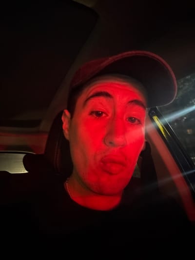 A man is posing for a selfie-style photo in what appears to be a vehicle at night, illuminated by a strong red light. The man is wearing a dark-colored baseball cap. He has a light complexion and his face is turned at an angle to the camera. His lips are pursed in a kissing or puckered expression. The background is predominantly dark, with vague shapes suggesting the car's interior and potentially the outside environment visible through the windows. The location is not discernible from the image.