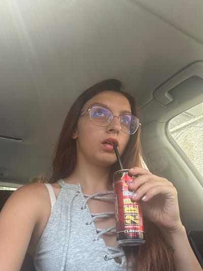 This is a medium shot, eye-level selfie taken inside a car, with a young woman holding an energy drink can to her lips and taking a sip. She is wearing large, gold-rimmed, square-shaped glasses and a gray, ribbed tank top with a lace-up neckline. Her hair is long and auburn, and it falls over her shoulders. The woman is looking to the right of the frame. The interior of the car is visible, including the ceiling, a handle, and the window. Outside the window, there is a blurred background of buildings and vegetation. There is a faint glare on the car's ceiling, indicating it is daytime. The image does not provide any specific geographical markers that would allow for its localization in Tryavna, Bulgaria.