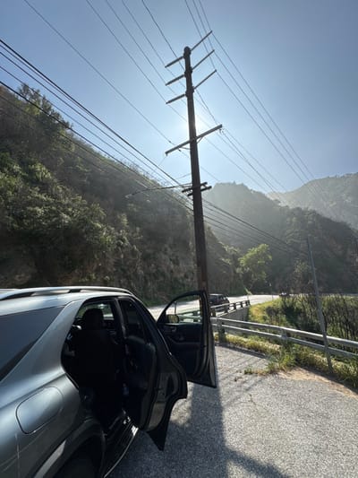 An overhead-lighted forensic image captures a dark gray or black SUV parked on the asphalt shoulder of a winding mountain road within the Unincorporated Santa Monica Mountains, United States of America. The vehicle's front and rear passenger doors are open, revealing dark interior seating. A tall wooden utility pole with multiple cross arms and numerous power lines dominates the midground, silhouetted against a bright, hazy sky with the sun positioned directly behind it. The road curves to the right, bordered by a metal guardrail. To the left, a steep, vegetated hillside rises. In the distance along the road, another dark vehicle is visible. The environment features verdant and partially fire-affected mountainous terrain under clear, sunny conditions. No individuals are visible.