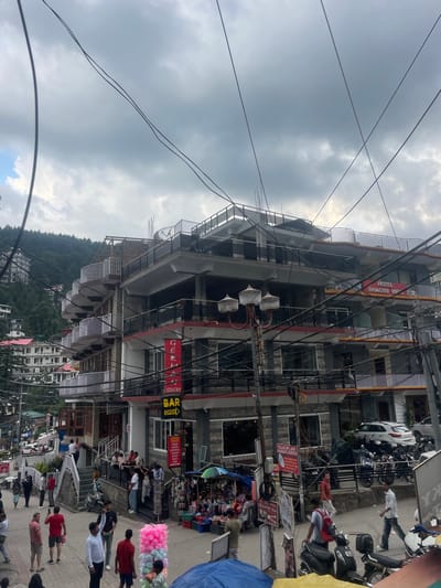 This media file presents a bustling street scene in Bhagsu, India, under an overcast sky during the daytime. The image, originally rotated, depicts a multi-story urban environment nestled against a backdrop of lush, tree-covered hills.

Numerous people are visible on the street, engaging in daily activities such as walking, standing, and congregating near shops. The street is lined with commercial buildings, many with balconies and a dense network of overhead power lines crisscrossing the sky. Several motorcycles and scooters are parked along the roadside, and a white car is also visible. Street vendors add to the activity, with colorful merchandise, including bright pink cotton candy, displayed prominently.

Notable details include the heavy infrastructure of power lines, typical of many Indian towns, and the blend of commercial and residential architecture. The weather is bright but predominantly cloudy, with occasional patches of lighter sky suggesting a break in the cloud cover.

Several signs and texts are visible, indicating the nature of the establishments:
*   "HOTEL BHAGSU VIP" is displayed on a prominent building.
*   "GERMAN BAKERY BAR OPEN" appears twice.
*   A detailed menu board for the German Bakery lists "INDIAN CHINESE CONTINENTAL MEXICAN ITALIAN ISRAELI FOOD."
*   "LAX SPA AND MASSAGE CENTER" is another clear sign.
*   "YES COMPORT VILLA" and a partial "YX" are also present.

The scene captures the vibrant, active atmosphere of a small town in a hilly region, characterized by commercial activity and everyday life.