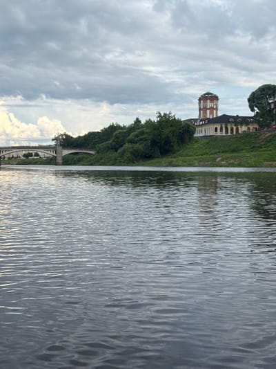 The image depicts a serene riverside scene with an old building and a bridge. The building, possibly a former industrial structure, features a prominent, octagonal brick tower with arched windows, set against a backdrop of a cream-colored main structure with dark dormer windows. Lush green trees line the riverbank, obscuring some of the building and leading down to a white arched bridge spanning the water. The river itself occupies the right side of the frame, its surface rippling gently, reflecting the overcast sky. In the distance, the sky is a mix of soft grey clouds and brighter patches, suggesting either early morning or late afternoon. The scene is quiet and tranquil, with no visible people or activity, evoking a sense of calm and historical reflection. The overall impression is of a picturesque and slightly melancholic landscape in Vologda, Russia, characterized by its historical architecture and natural beauty.