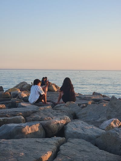 A photograph shows a group of four people sitting on a rocky shoreline along the coast of Quarteira, Portugal, during sunset. The sky exhibits a gradient of light blue to soft orange, indicating the sun is low on the horizon. The sea is calm with gentle ripples, and a few small boats are visible in the distance.
In the foreground, a person in a striped t-shirt and dark shorts is seated on a large rock, looking down at a mobile phone. To their right and slightly behind, another person with dark hair, wearing a black t-shirt and shorts, is also seated facing the sea. Further back, two younger individuals are positioned on rocks near the water's edge. One of them, wearing a blue shirt, is holding a mobile phone.
The rocks are large and weathered, forming a natural breakwater. The lighting suggests it is late afternoon or early evening, with the sun casting long shadows and illuminating parts of the rocks with a warm, golden hue. The overall atmosphere is serene and relaxed, with the individuals seemingly enjoying the coastal view and the quiet evening.