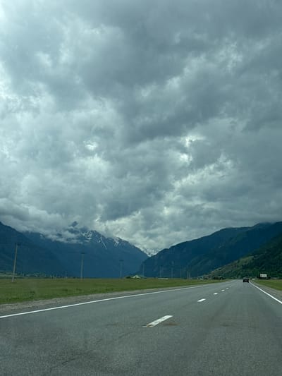 The image depicts a view from a vehicle traveling on a paved road through a mountainous, rural landscape. The road has white lane markings and appears to be a two-lane highway. To the left of the road is a grassy field. In the distance, a range of dark, verdant mountains rises, with some snow-capped peaks visible towards the center. The sky is overcast with heavy, gray clouds. Several utility poles are present along the side of the road. A dark-colored vehicle is visible in the distance on the right lane of the road. A single, small building with a green roof is situated in the middle distance on the left side, nestled amongst trees at the base of the mountains. The scene appears to be located in or near Верхняя Теберда, Russia, given the mountainous terrain and the presence of utility poles, which are common in such regions.