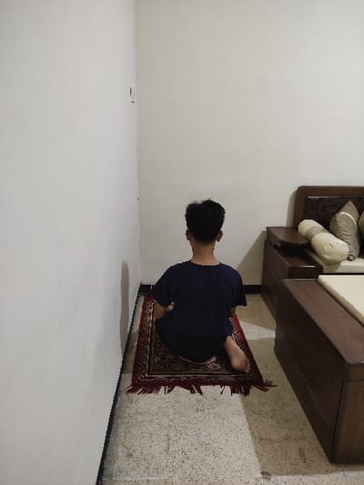 The image shows a person sitting on a prayer rug in the corner of a room. The person is wearing a dark blue t-shirt and has short black hair. The person is sitting with their legs crossed and their back straight. The rug is red with gold accents. The floor is tiled in a light beige color. There is a white wall to the person’s left and a wooden bed to the right. The bed has a white mattress and a brown wooden frame. There are two pillows on the bed. The room is lit by overhead lighting. The location, based on the coordinates, is in the city of Kecamatan Lawang, Indonesia. The coordinates provided place the location in a residential area of Lawang. The scene is set indoors with the person engaged in prayer.