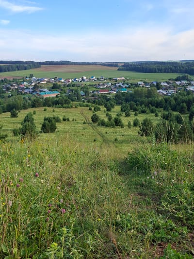 The image depicts a panoramic view of a rural village nestled amidst rolling green hills under a bright blue sky with scattered white clouds. The village, identifiable as being in Chumnoye, Russia, features a collection of houses with varied roof colors, including red, blue, and grey, surrounded by lush trees. A dirt path winds through the grassy fields, leading towards the village from the foreground, which is covered in dense wildflowers and tall grass. In the distance, agricultural fields and forests create a layered landscape extending to the horizon. The scene is bathed in natural daylight, suggesting a clear day. There are no people visible in the image.