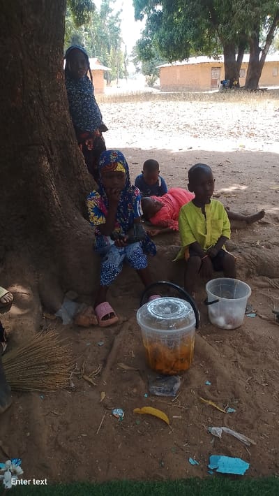 The image depicts an outdoor scene in Rinze, Nigeria, under a large tree providing shade. The ground is dry, light brown earth with scattered debris.
Five children are present in the foreground and mid-ground.
* A young female, partially obscured by the tree trunk on the left, wears a blue patterned head covering and dark, patterned attire. She looks towards the viewer.
* Another young female is seated on the tree's exposed roots, wearing a blue, yellow, and red patterned head covering, a blue patterned top and trousers, and pink slip-on sandals. Her right hand is near her mouth, while her left hand holds a small dark object. She looks forward.
* Immediately to her right, a young male sits, wearing a solid light blue top, looking downwards.
* To the right of this male, a child in a pink or red garment lies prone on the dusty ground, with their face obscured.
* Further to the right, a young male sits, wearing a lime green short-sleeved tunic and dark shorts, looking towards the right.
In the foreground, on the ground, are a large clear plastic cylindrical container with a lid, filled with orange and brown food items, and a smaller clear plastic bucket with a handle and lid, containing several white ovoid objects. A straw broom lies on the ground near the tree roots. Scattered litter, including a yellow leaf and blue plastic fragments, is visible on the dusty surface.
The background features several single-story buildings of light brown construction, likely mud brick, with light-colored roofs. Dry vegetation and additional trees are visible in the distance under a bright, sunny sky. Two individuals are discernible seated under a tree near one of the distant buildings on the right.