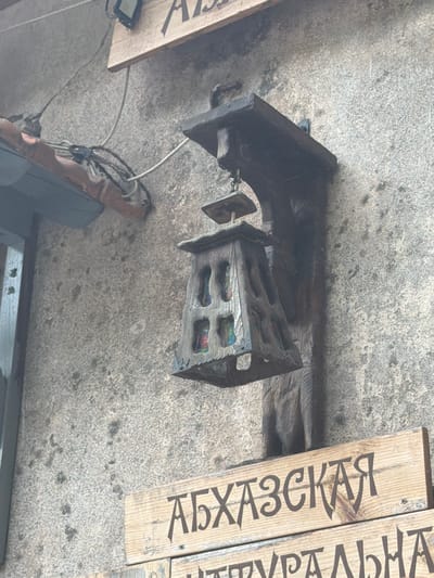 A rustic, carved wooden lamp hangs from a weathered stone wall. The lamp features an ornate, cage-like design with what appears to be stained glass panels, though their colors are muted. Above the lamp, a wooden beam supports it, with visible electrical wires running along the wall and a small, black electrical box. Below the lamp, two wooden signs with Cyrillic lettering are affixed to the wall. The top sign reads "АБХАЗСКАЯ" and the bottom sign is partially visible, with the letters "ТУРАЛЬНА" appearing. The wall itself is textured and speckled, indicating an outdoor or an old building facade, consistent with the historical architecture found in cities like New Athos, Abkhazia, a location known for its ancient monasteries and natural beauty. The overall scene evokes a sense of old-world charm and historical preservation.