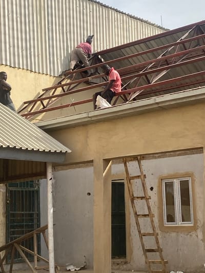 The image captures an active construction site in Bisichi, Nigeria, under daylight conditions. Three adult males are present around a building under construction or renovation. In the upper center, a man wearing a pink long-sleeved shirt, light trousers, and a striped head covering is on his hands and knees on a reddish-brown metal roof truss structure. To his right, another man, dressed in a pink short-sleeved shirt and dark shorts, is seated on a horizontal beam of the roof truss, holding a white bag on his lap. A third man, wearing a dark sleeveless shirt, stands on a lower, grey corrugated metal roof in the far left, observing the scene with his arms crossed. The building features a freshly rendered light-brown upper wall. Below this, a lighter, unfinished wall section contains a newly installed white-framed window with multiple panes and a dark door within an open doorway. A wooden ladder, constructed from rough timber, leans against this unfinished wall section. Behind the primary roof truss, a large, grey corrugated metal wall or roof of an adjacent structure is visible. Small pieces of white debris are scattered on the ground below the construction area.