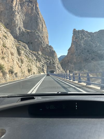 The image is taken from the perspective of someone driving a car on a highway that cuts through a narrow mountain pass. On the left and right sides of the road, there are steep, rocky cliffs with sparse vegetation. The road itself is paved, with clear lane markings and a white barrier. In the distance, the road leads into a tunnel carved into the mountainside. A blue metal railing runs along the right side of the road, separating it from a lower level. The sky is clear and blue. The car's dashboard is visible in the foreground, showing a digital display that indicates a speed of 66. The scene appears to be located in the mountainous region surrounding Altea, Spain, possibly on a route that connects to tunnels and passes in the area.