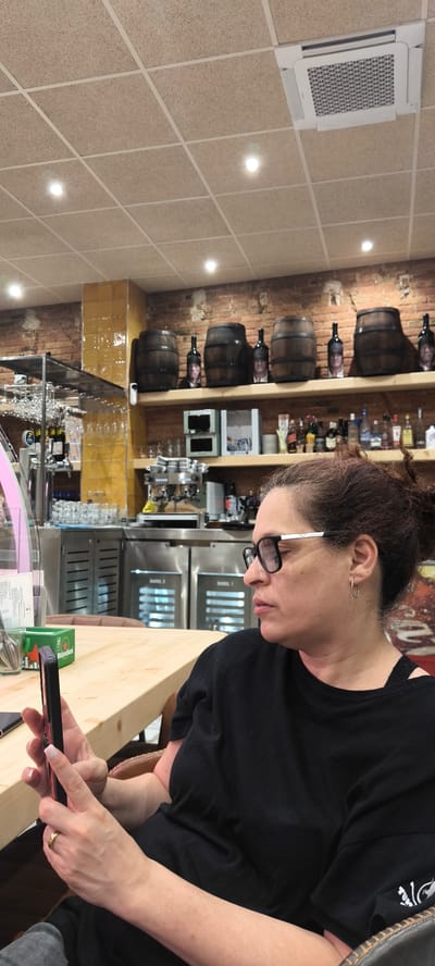 A woman with curly brown hair is seated at a wooden table inside a restaurant. She is wearing black-rimmed glasses, a black t-shirt, and hoop earrings, and she is looking at and holding a cell phone. The restaurant's interior features a textured tile ceiling with recessed lighting and a white air conditioning unit. A brick wall with a yellow tiled section is visible, with a shelf holding wooden barrels with human head cutouts, bottles, and glasses. Below the shelf, stainless steel equipment, including an espresso machine and refrigerators, is seen. Wine glasses hang from a rack above the bar area. A green Heineken box sits on the table, next to the woman. The city is Ripollet, Spain.
