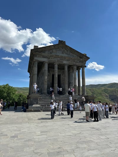 The image captures the Garni Temple, an ancient Greco-Roman colonnaded temple located in Garni, Armenia. The temple, constructed from basalt, stands on a raised platform with a wide staircase leading up to it. A group of tourists are gathered on the stairs and in front of the temple, many of them dressed in casual summer attire. The sky is a vibrant blue with scattered white clouds, and the surrounding landscape consists of rolling green hills. The scene is bathed in bright sunlight, casting distinct shadows on the stone paving.