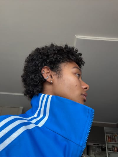 A young person, with dark curly hair and visible earrings, is shown in profile against a grey wall. They are wearing a bright blue jacket with white stripes on the shoulder and collar area. The person's face is partially obscured by the angle, but their profile, including their nose, lips, and ear, is visible. The jacket appears to be a track jacket, with a zipper partially visible. The background suggests an indoor setting, possibly a home or studio, with a glimpse of kitchen shelving on the right side of the image. No specific location within Champs-sur-Marne, France can be determined from the visual information provided.