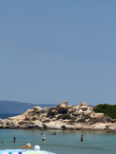 The media file shows a rocky coastline with clear blue water. Several people are visible in the water, some swimming and others relaxing on inflatable rafts. The rocky formation on the left side of the image is a prominent feature, with lush green vegetation growing at its top. The sky is a clear, pale blue, suggesting a sunny day. The scene appears to be a tranquil beach or cove in Greece, likely Sithonia, as indicated by the location context. The presence of people enjoying the water and the calm weather contribute to a relaxed and peaceful atmosphere. There are no visible text elements in the media file.