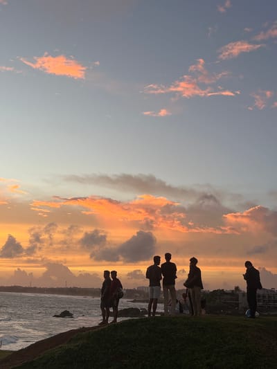 The image depicts a group of individuals silhouetted against a vibrant sunset sky, likely taken in Galle, Sri Lanka. The sky is filled with dramatic clouds painted in shades of orange, pink, and grey, suggesting it's either sunrise or sunset. A few wisps of orange clouds are scattered across the pale blue sky, with larger formations of pink and grey clouds dominating the horizon.
In the foreground, a line of people is standing on a grassy embankment, looking out towards the water. They are all in silhouette, making it difficult to discern individual features, but their postures suggest a relaxed, observational mood. Some are standing together, possibly friends or family, while others are more spread out. The water appears to be calm, with a few rocks visible at the edge of the embankment.
Beyond the water, the distant coastline is visible, with buildings and possibly a lighthouse or tower. The overall impression is one of a serene evening or early morning scene, bathed in the warm glow of the setting or rising sun. The rich colors of the sky create a visually striking backdrop for the silhouettes of the people and the natural landscape. There is no visible text in the image.