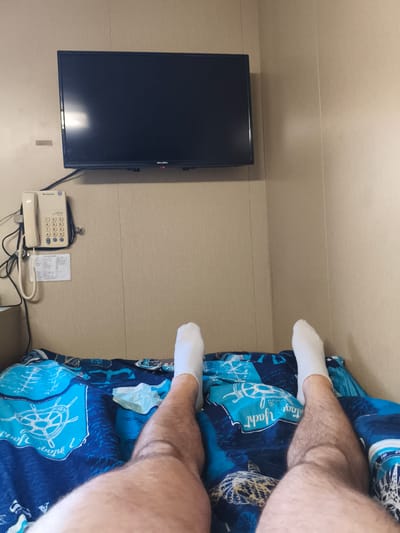 A person's legs and feet, clad in white socks, are resting on a bed with blue bedding featuring a nautical theme. A black television is mounted on a beige, paneled wall above a cream-colored telephone. The scene appears to be an indoor space, possibly a cabin or small room. There is no discernible indication of the location being Velsen-Noord, Netherlands.