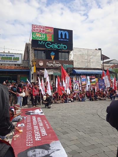 The image shows a gathering of people in front of a building with a "My Gelato" sign in Yogyakarta, Indonesia. There are a large number of people, most of whom are wearing red clothing, and they are holding banners with various messages written in Indonesian. The banners feature slogans like "Federasi Serikat Pekerja" and "Persatuan, Perjuangan Sambut Hari Nelayan." Some people are standing while others are sitting, and there is a banner stretched out on the ground in front of them. The building in the background has a sign for "Mimicucu" and "Salad Nyoo." There is a large, blank billboard on the side of the building. The scene is in front of a storefront. It is a bright day, with a blue sky and some clouds in the sky. The people are likely participating in a demonstration or rally. The event is taking place on a street with a brick sidewalk.