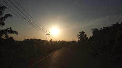 This is a horizontal, eye-level, medium shot of a paved road at sunset in Seuneubok, Indonesia. The sun is a bright, large circle of light in the center of the image, casting a warm glow across the hazy sky. Power lines stretch across the upper portion of the frame, supported by wooden poles that are silhouetted against the bright sky. The road is empty except for a single motorbike visible in the distance on the right side of the frame. The surrounding landscape is dense with tropical vegetation, including palm trees, which are also silhouetted against the sky. The overall mood of the image is serene and peaceful, with the warm light of the setting sun creating a calming atmosphere.