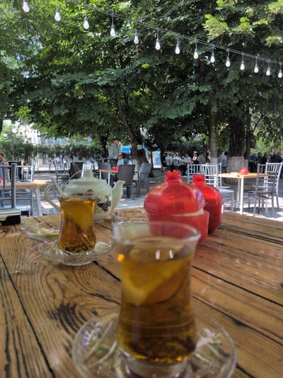 The image depicts an outdoor seating area of a cafe or restaurant, likely located in Şəki, Azerbaijan, given the presence of traditional tea glasses. In the foreground, a rustic wooden table is set with two glasses of tea, one filled with amber liquid and a lemon slice, and another partially visible glass. A white teapot adorned with a butterfly motif sits between the glasses. Two bright red, pomegranate-shaped sugar containers are also on the table. In the background, several trees provide shade, with string lights hung between them. People are seated at tables in the mid-ground, some conversing, while others are engaged with their drinks. The setting appears relaxed and casual, suggesting a pleasant day at an outdoor eatery.