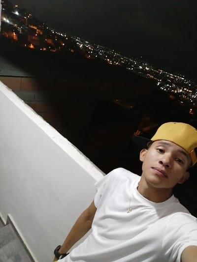 A young man in a white t-shirt and yellow baseball cap poses for a selfie at night. He is wearing a necklace. The background shows a city with numerous lights, possibly from a high vantage point like a balcony or rooftop. A light-colored wall is visible to the left, and there is a sense of elevation in the scene. The man's gaze is directed towards the camera.