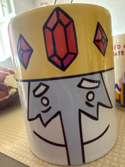 The image shows a close-up of a white mug with a yellow band at the top, decorated with red and pink geometric gems. The mug also features a simplified cartoon face with light blue skin, black outlines for eyes, nose, and mouth, and a jagged black line across the forehead. The background is blurred but appears to be an indoor setting with a patterned surface. To the right of the mug, another mug is partially visible, with text that includes the words "ED A" and "RATE". The mug is placed on a light-colored, textured surface. This scene does not have any relation to Biysk, Russia.