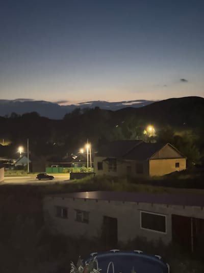 This image depicts a twilight scene in the city of Mednogorsk, Russia. The sky is a gradient of deep blue at the top, fading to a lighter, yellowish hue near the horizon, with faint clouds visible. In the middle ground, a rural village landscape is illuminated by streetlights. Several houses with pitched roofs are visible, some with lights on inside. A dark-colored car is driving on a paved road in front of one of the houses. Streetlights cast a warm glow on the surrounding area, illuminating trees and other vegetation. In the background, dark, silhouetted hills or mountains rise against the twilight sky. In the foreground, a part of a building with windows is visible, and further down, a blue inflatable pool is partially seen. The overall atmosphere is calm and serene, characteristic of a rural evening.