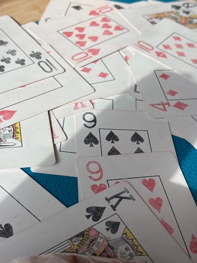 The scene is located in Ivanovka, Russia. The image provides a close-up, high-angle view of a pile of scattered and overlapping playing cards resting on a textured, teal-colored surface. The surface exhibits a consistent, fine-grain pattern, possibly a tabletop or mat. Illumination from the upper-left casts pronounced shadows across the cards, accentuating their contours and the layered arrangement. The cards are standard playing cards, primarily white-backed with red (hearts and diamonds) and black (spades and clubs) suits. Visible cards include a '10' of Clubs (upper left), a '10' of Hearts (upper center-left), a partially visible '10' of Diamonds (mid-left), a '4' of Diamonds (upper right), a '9' of Spades (mid-right), a '9' of Hearts (lower-center), a King of Spades (lower-left-center), and a partial Queen of Hearts (lower-left). The cards display minor wear and scuffing consistent with regular use. No individuals, actions, or interactions are depicted; the objects are static.