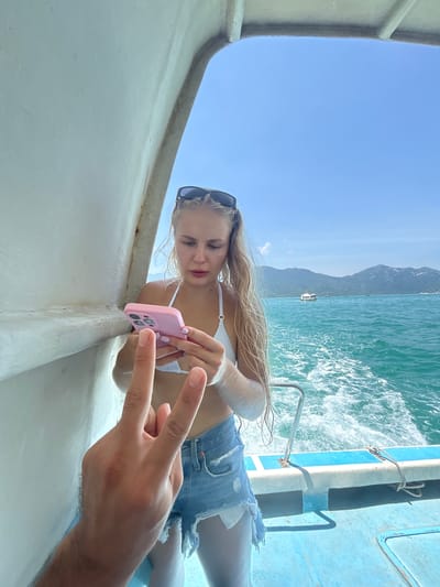 A woman wearing a white bikini top and denim shorts stands on the deck of a boat in Nha Trang, Vietnam. She is holding a pink smartphone and looking at it. Her hair is long and blonde, and she is wearing sunglasses on her head. A man's hand is visible in the foreground, holding up two fingers in a peace sign. The boat is on the water, with waves and a distant coastline with mountains in the background. The sky is clear and blue.