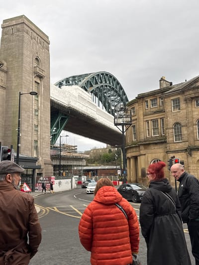 A street scene features the iconic Tyne Bridge in Newcastle upon Tyne, with scaffolding partially obscuring its lower structure. Pedestrians in varying colored winter coats populate the foreground. A tall stone building stands to the left, while another, more ornate building with multiple windows, is visible in the background. Traffic signals are present, and cars are seen on the road between the buildings. The sky is overcast.
