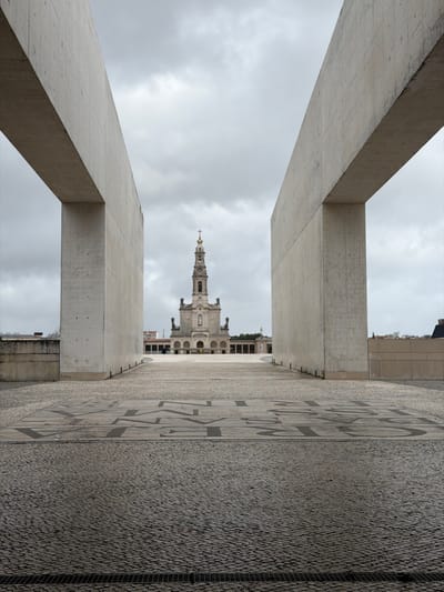 The image depicts an exterior view within the Sanctuary of Fátima, Portugal, looking directly towards the Basilica of Our Lady of the Rosary. The scene is framed by two large, raw concrete vertical structures, possibly architectural elements of the modern Basilica of the Holy Trinity or an adjacent colonnade, creating a narrow visual corridor.
The foreground consists of dark grey cobblestone paving (calçada portuguesa) with a horizontal metal drainage grate at the very bottom. Immediately beyond this, a lighter-colored section of pavement features intricate, dark geometric patterns and possibly stylized text. This paving extends into a vast, open plaza leading towards the basilica.
In the midground, the Basilica of Our Lady of the Rosary stands centrally. It is a light-colored, classical-style building featuring a prominent, tall bell tower (campanile) topped with a golden cross and an onion dome. The basilica's facade includes arched entrances, windows, and sculptural details. Large statues flank the main body of the basilica on either side, and colonnaded walkways extend horizontally from its base.
The plaza appears largely empty, with only a few indistinct, small figures visible far in the distance near the basilica. The sky is uniformly overcast with grey clouds. No significant interactions or specific actions are discernible. The overall environment is one of expansive, solemn architectural space under a muted sky.
