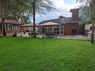 The image shows a grassy courtyard located at coordinates -0.754, 36.437 in Karagita, Kenya.  The courtyard features a red brick building with a tiled roof and a large fireplace on the back wall. The building is surrounded by a stone patio and two beige canopies that are supported by four metal poles.  Beneath each canopy are two tables with four chairs each, and a third table with four chairs is positioned between the two canopies.  The courtyard is also surrounded by trees and foliage. There are no people in the image.  The image was taken during the day. The sky is partly cloudy, with blue sky visible through the clouds. The ground is covered in green grass.  The image was taken from a low angle, looking towards the building. The image is in focus and well lit.