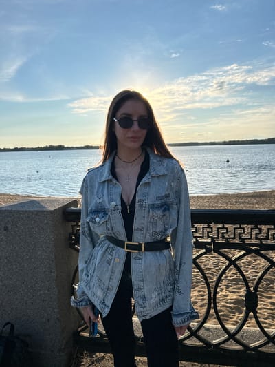 The image shows a young woman standing by a body of water with a beach and a decorative metal fence behind her. The sun is setting, casting a warm glow and creating a silhouette effect. The woman is wearing a denim jacket over a dark top, a black belt, and black pants. She has sunglasses on and is wearing necklaces. The background includes a wide body of water, a sandy beach, and trees in the distance. The location is identified as Novokuybyshevsk, Russia, likely along the Volga River, as this is a prominent geographical feature near the city. The overall impression is one of a casual, relaxed outdoor scene during sunset.