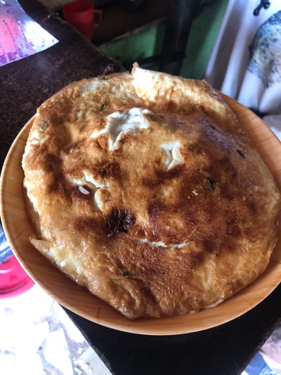 A close-up, overhead shot shows a golden-brown omelet on a light brown, circular plate. The omelet is irregularly shaped with a slightly raised, puffy center that has lighter, cream-colored sections, indicating undercooked egg. The edges of the omelet are darker and crisper. Scattered within the omelet are small green specks, possibly herbs, and tiny flecks of orange, possibly spices or dried chilies. The plate rests on a dark, textured surface, likely a wooden table. In the upper left corner, partially visible, is a clear plastic bag with purple text. Behind the plate and to the left, a red mug is visible. The background is out of focus, with hints of green and white fabric. There are no people or discernible actions taking place. The scene appears to be indoors, likely in a casual dining setting. The image does not contain any specific visual information to determine its location within the city of Kabong, Nigeria.