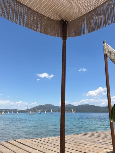 The image shows a close-up view of a wooden pier leading to a body of water, likely the Ionian Sea. In the foreground, the pier is constructed of weathered wooden planks. Two wooden poles, part of a beach umbrella or shade structure, are visible. The left pole is closer and in focus, with the right pole further away and slightly out of focus. Both poles have decorative fringe hanging from them. The water is a clear turquoise color, with gentle ripples. A fleet of sailboats is dotted across the water in the middle ground. Beyond the water, a chain of lush green, mountainous islands forms the horizon. The sky is a vibrant blue with scattered white cumulus clouds. The scene suggests a serene beach or waterfront location within the Municipal Unit of Lefkada, Greece, known for its beautiful islands and coastal scenery. There are no people visible in the image.