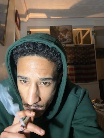 A man wearing a green hooded sweatshirt is holding a lit marijuana cigarette. Smoke is emanating from the tip of the cigarette. Behind him is an American flag and framed artwork hanging on a wall. The location is likely indoors, but the specific city and country details of Tanger, Morocco are not verifiable from the image.
