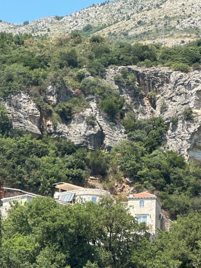 The image depicts a scenic view of Perazića Do, Montenegro, focusing on a rocky mountainside covered in lush green trees. In the foreground, partially obscured by the foliage, are two stone buildings. The building on the left appears to be a complex with a flat roof and some covered outdoor areas. To its right, a more distinct structure stands with a red-tiled roof, two stories, and visible windows. The mountain slopes behind the buildings are characterized by rugged rock formations and dense vegetation, with a clear blue sky overhead. The overall scene suggests a tranquil, natural environment with human habitation integrated into the landscape.