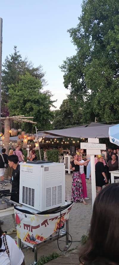 The image depicts an outdoor gathering at a beachfront or sandy venue, likely a bar or restaurant, under a bright sky. In the foreground, a white ice machine with visible vents sits on a wheeled cart, partially covered by a fabric adorned with images of waffles and fruit. To the left of the machine, a person wearing a black shirt and apron stands near a grilling or cooking apparatus.
Further back, several people are visible mingling. Two women in the center, one wearing a black top and a floral skirt and the other in a white top, are posing together, both wearing gold party hats. Behind them, a white sign with "#GLAUBAENDIG" is visible. The venue features outdoor seating, with tables and chairs, and is partially covered by large white canopies. String lights are strung across the area, suggesting it is an evening event or has been set up for nighttime enjoyment. Trees with lush green foliage form a backdrop, and hanging pendant lights are visible in the mid-ground. The overall scene suggests a lively social event in a relaxed, outdoor setting. The provided context mentions Linz, Austria, which suggests this location is within or near the city.