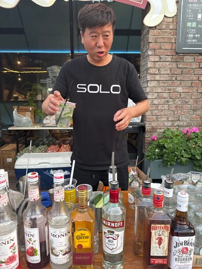 In the foreground, a man wearing a black t-shirt with "SOLO" printed on it is standing behind a table laden with bottles of alcohol and syrups. He is holding a clear plastic cup with a drink, a straw, and ice. In front of him are several bottles, including Jose Cuervo Especial Tequila, Smirnoff No. 21 Vodka, Beefeater London Dry Gin, and Jim Beam Bourbon. There are also bottles of Monin syrups, such as Rose and Coconut flavored syrups. The background shows a brick wall, potted plants with pink flowers, and what appears to be a cooler filled with bottles. In the middle ground, a table holds more bottles and drinks. In the far background, there are shelves with more items and a white cooler. The overall scene suggests an outdoor bar or beverage stall, possibly in a commercial area. The sign on the brick wall is partially visible and written in Chinese characters. The location is identified as Chaoyang District, China.