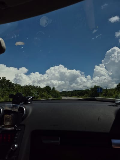 The image depicts a driver's perspective from within a vehicle, looking forward onto a two-lane asphalt road curving through a rural landscape under a bright blue sky with large, white cumulus clouds. Dense green deciduous trees and bushes line both sides of the road, extending into the distance. On the right side of the road, a blue rectangular road sign with three upward-pointing white arrows indicates traffic lane information. Faintly visible in the distant left are structures resembling electricity transmission towers above the tree line. The vehicle's dark-colored dashboard occupies the lower portion of the frame, showing a black suction-cup phone mount in the center-left, circular air vents, and part of a control panel with red illuminated elements on the lower left. Reflections on the windshield include a white disposable cup in the upper right quadrant and other indistinct shapes. The word "AIRBAG" is faintly visible on the passenger side of the dashboard. This scene is situated in Bogyovtsi, Bulgaria.
