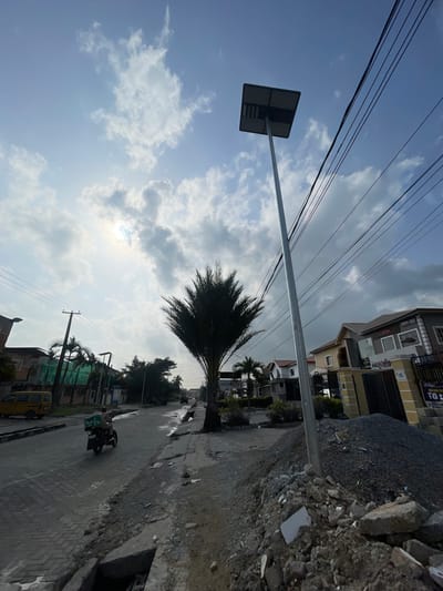 The scene captures a street in Moba, Nigeria. The road is paved with bricks and lined with buildings of various styles, including multi-story residential structures. Construction debris is piled on the right side of the frame, bordering the road. A palm tree stands prominently near the roadside. Electrical wires span overhead. A motorbike with a rider is visible on the street. A street sign is positioned on the left, reaching across the road. The sky is partly cloudy, with glimpses of sunlight.