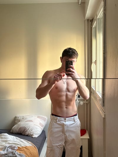 The image displays a male individual capturing a mirror selfie inside a room within a building located in Rennes, France. The subject, appearing to be a young adult, has short, dark hair and a muscular physique, evident in his defined chest and abdomen. He is shirtless, revealing a silver-colored cross necklace around his neck and an earring in his left ear. He wears light-colored cargo pants, with a dark waistband featuring a red stripe visible above them. His face is obscured by the dark-cased smartphone he holds in his right hand, angled towards the mirror. His left hand is partially raised.
The room's environment consists of light-colored walls, illuminated by natural light entering through a white-framed, multi-pane window located on the right side of the frame. A vertical shadow line from this light source is visible on the wall. Below the window, a white radiator with a control knob is present. A small white table stands next to the radiator, holding a red, heart-shaped object. On the left side of the image, a bed with a dark mattress base, a light-colored pillow featuring a subtle geometric pattern, and rumpled orange and white bedding is partially visible.