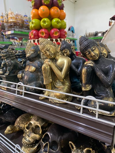 A horizontally-oriented close-up shot taken indoors shows rows of merchandise on shelves in a store. The location is Nusa Dua, Indonesia.
The items are on a white wire rack with a brown border. The top shelf has stacked, reclining Buddha statues. The next shelf contains skulls and turtles. To the left, behind the wire rack, is a display of produce, including oranges, green apples, and red apples. Other items in the background are small tourist items.
The Buddha statues are stacked. The top Buddha is black with gold accents, while the second Buddha is gold. The heads of the Buddhas have small tightly formed curls.
The skulls have large eye sockets and teeth. The turtle's shells are detailed with patterns.