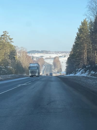 This image depicts a winter daytime scene on an asphalt highway in the vicinity of Сундур, Russia. The sky is clear and light blue. The road, appearing damp or partially melted, extends into the distance over undulating terrain.
In the foreground, a light-colored semi-truck with a cab-over-engine design and a box trailer is driving towards the viewer in the left lane, its headlights illuminated. Further down the road, two smaller passenger vehicles are visible, traveling away from the viewer.
The left side of the highway is bordered by a dense stand of coniferous trees, beyond which lie snow-covered fields, bare deciduous trees, and distant hills with scattered trees and structures indicative of a rural settlement. The right side of the road features a snow-covered embankment leading to a dense coniferous forest. All visible ground areas, excluding the cleared asphalt, are covered in snow, consistent with winter conditions. No people are discernible in the scene.